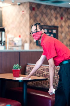 Waiter wearing a face mask cleans a table in a restaurant, maintaining hygiene standards.